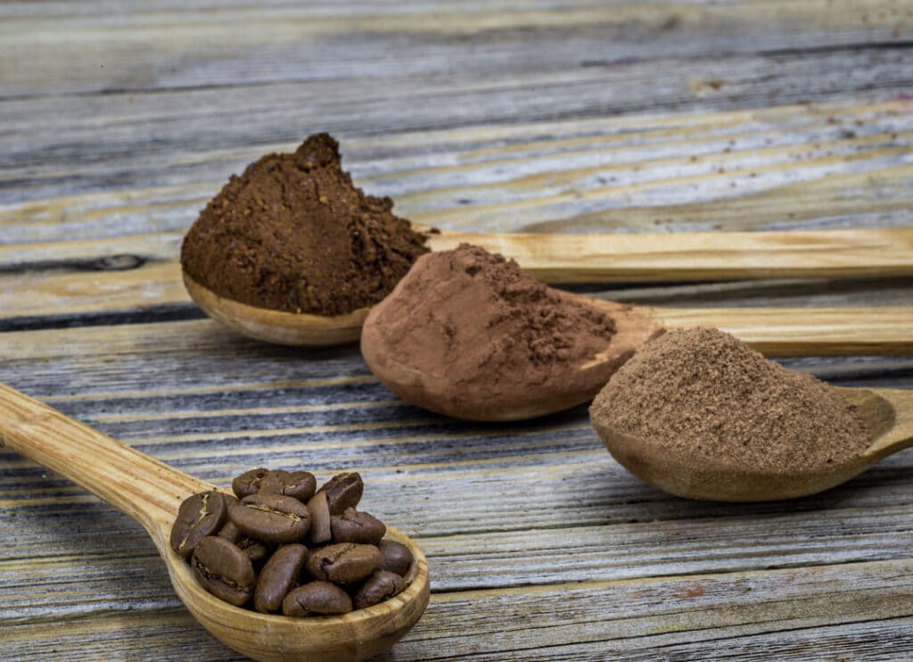 Wooden spoons with defatted cocoa powder, roasted cocoa beans, and different cocoa textures on a rustic wooden table, illustrating stages of cocoa processing.
