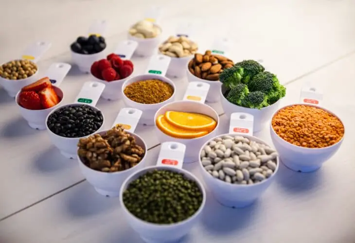 Alimentación plant based: tazas medidoras con legumbres, frutos secos, frutas y brócoli ordenados sobre una mesa de madera.